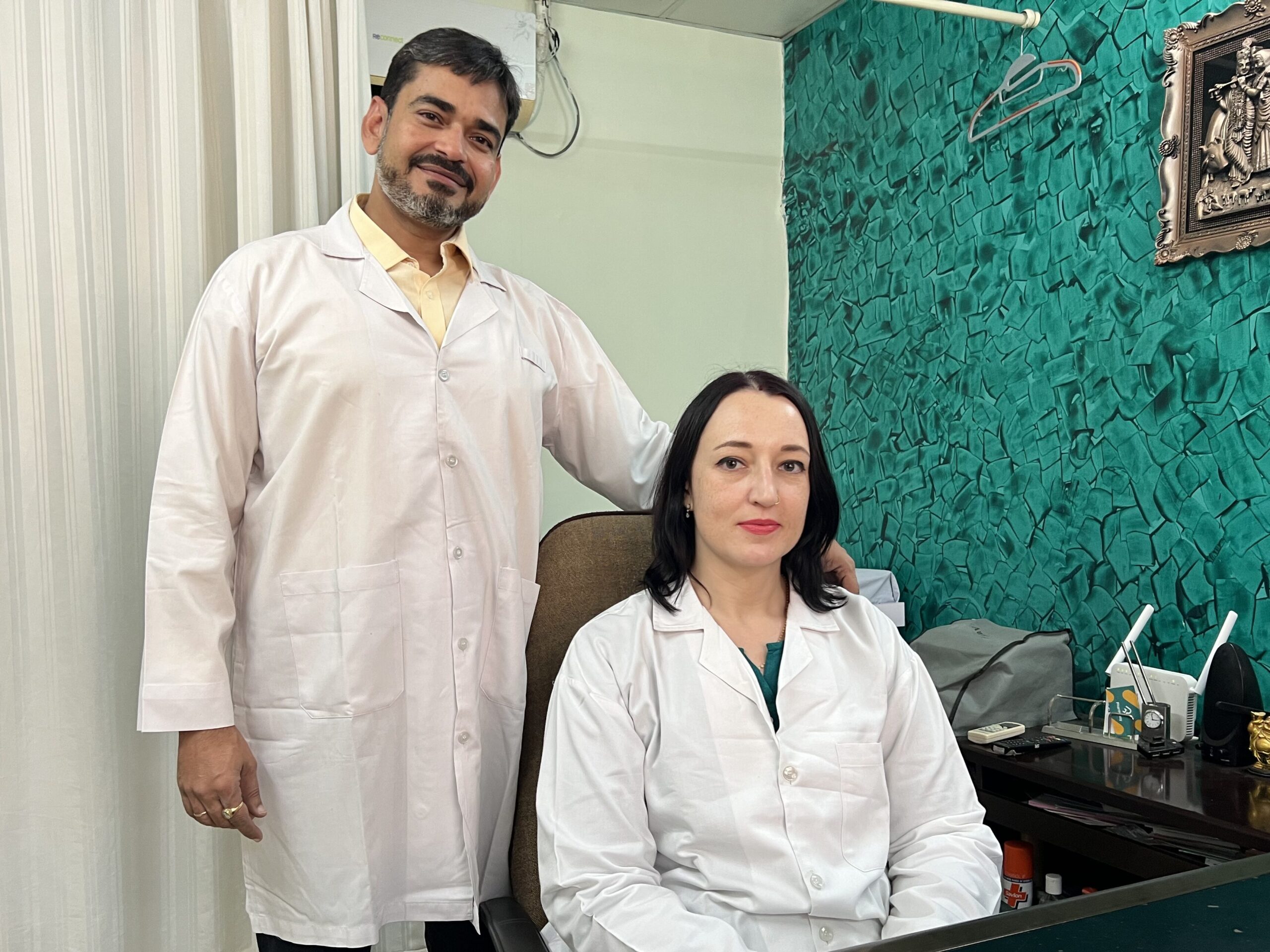 A male and a female healthcare professional in white lab coats are posing for a photo in an office setting. The male is standing behind the seated female, both of whom are smiling. The office has a green textured wall and various items on a desk.